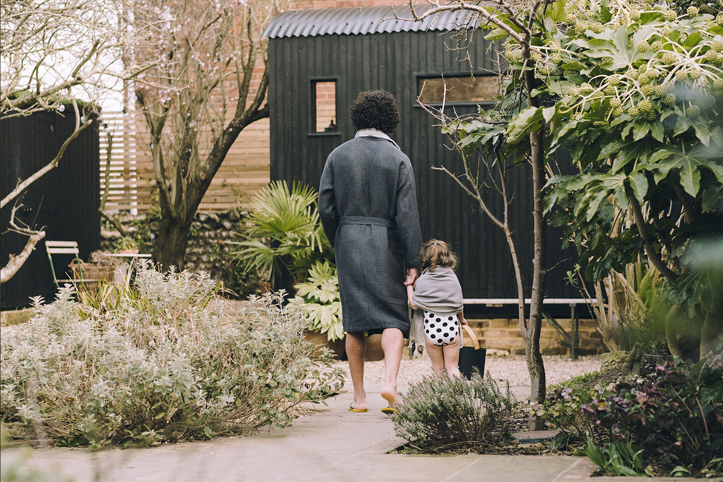 Man in dark grey dressing gown holding hands with small girl in black and white polka dot swimming costume and towel walking down a garden path towards a black sauna hut surrounded by trees